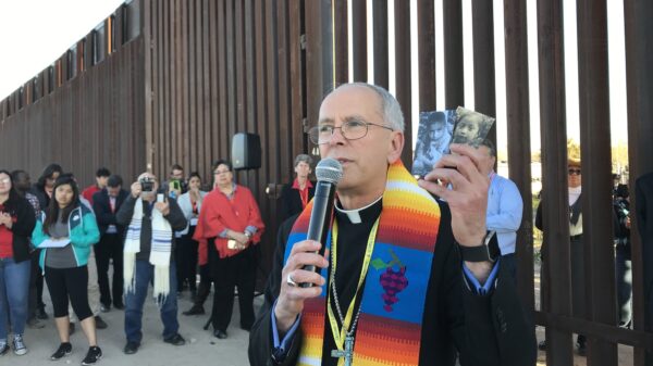 El obispo Mark J. Seitz de El Paso, Texas, es visto el 26 de febrero de 2019, en el muro fronterizo entre Estados Unidos y México sosteniendo fotos de dos niños migrantes que murieron bajo custodia estadounidense. (Foto CNS/David Agren)